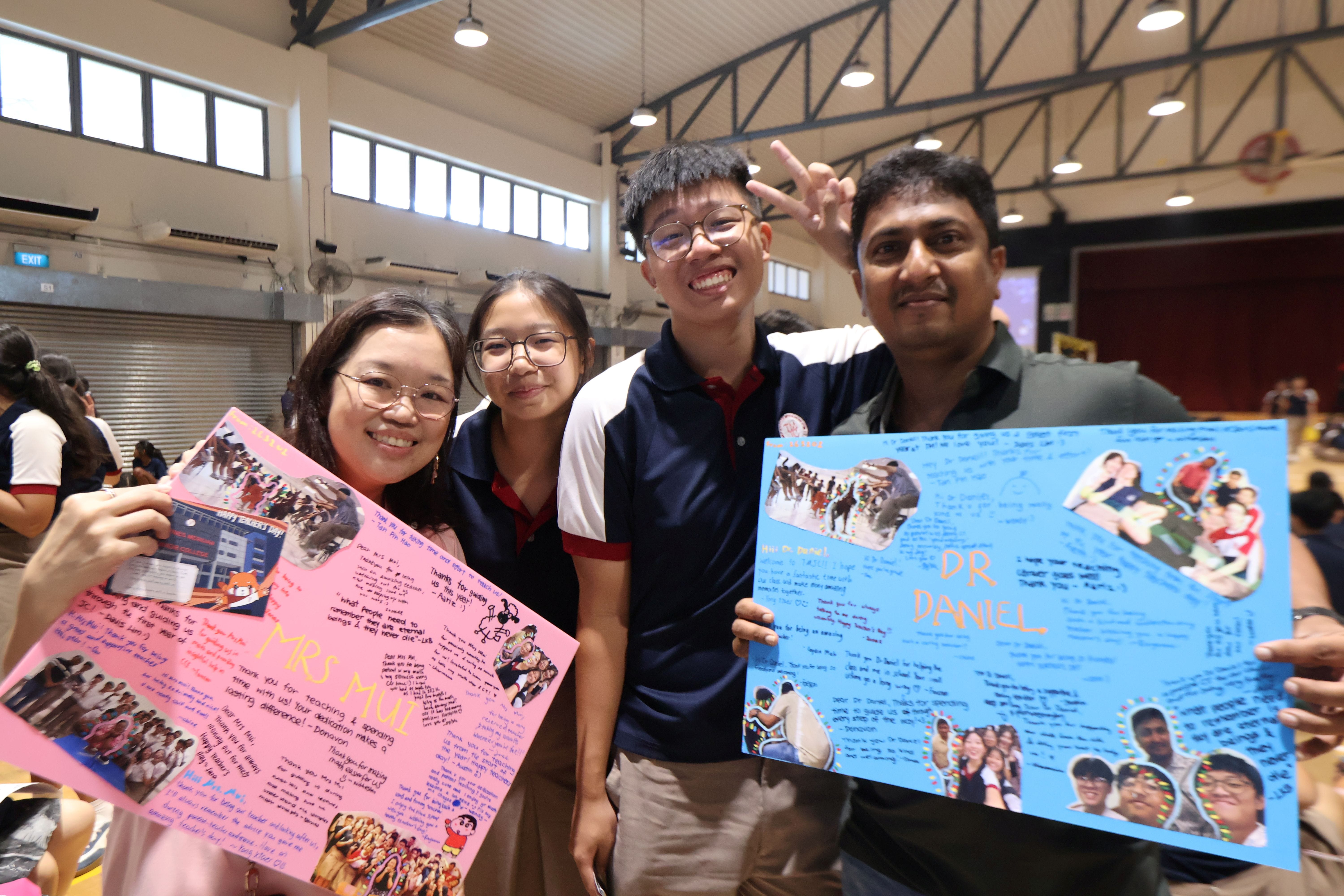 Four people — two teachers and two students — smile at the camera in a school hall, holding up large handmade appreciation cards covered in photos, messages, and drawings, addressed to "Mrs Mui" and "Dr Daniel."